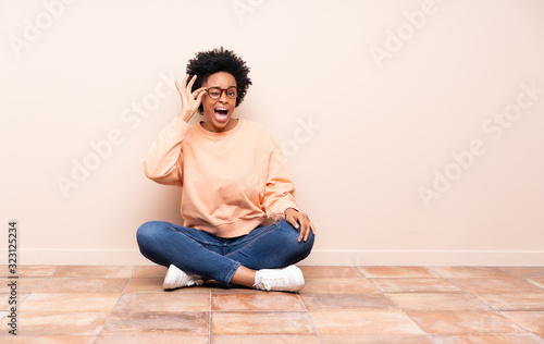 African american woman sitting on the floor with glasses and surprised © luismolinero