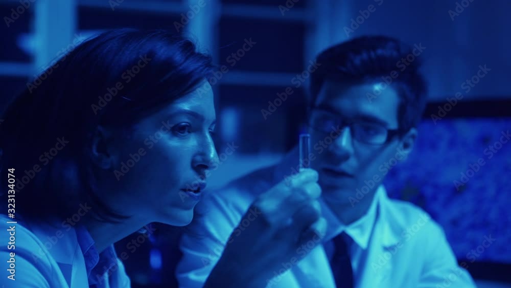 Blue toned monochrome shot of two shocked biochemistry scientists, man and woman in lab coats, examining carefully and discussing liquid in test tube in laboratory making scientific discovery Blue toned monochrome shot of two shocked biochemistry scientists, man and woman in lab coats, examining carefully and discussing liquid in test tube in laboratory making scientific discovery