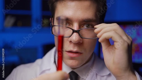 Zoom in closeup of young chemistry scientist examining red liquid in test tube and taking off eyeglasses to explore surprising experiment results looking shocked in laboratory