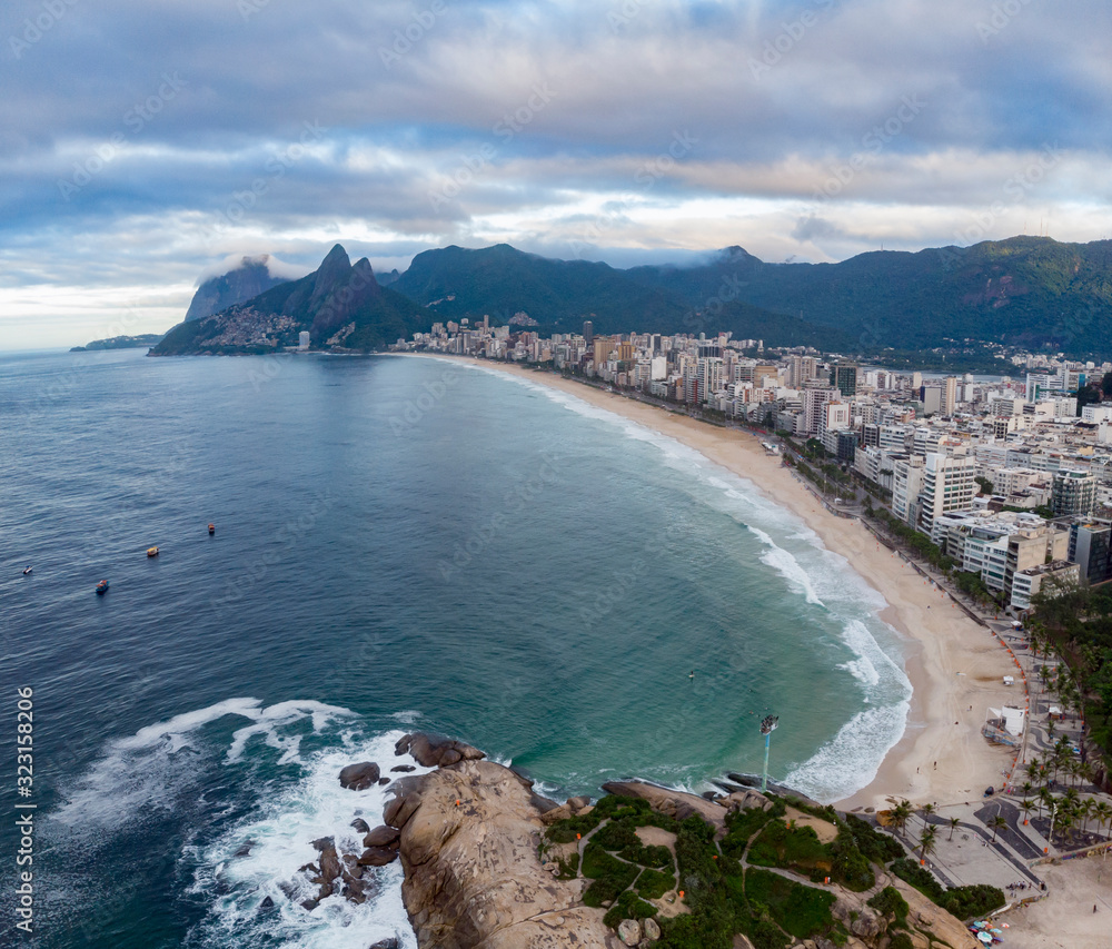 Arpoador rock in Rio de Janeiro with Ipanema beach in the foreground and wider cityscape including Two Brothers mountain in the background against an overcast sky at sunrise. Aerial seascape.