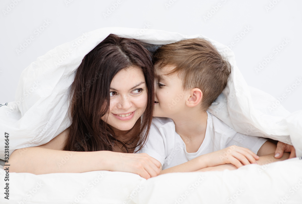 Mom and son are lying under the covers at home in the bedroom, the son whispers something in the mother’s ear Mom and son are lying under the covers at home in the bedroom, the son whispers something in the mother’s ear