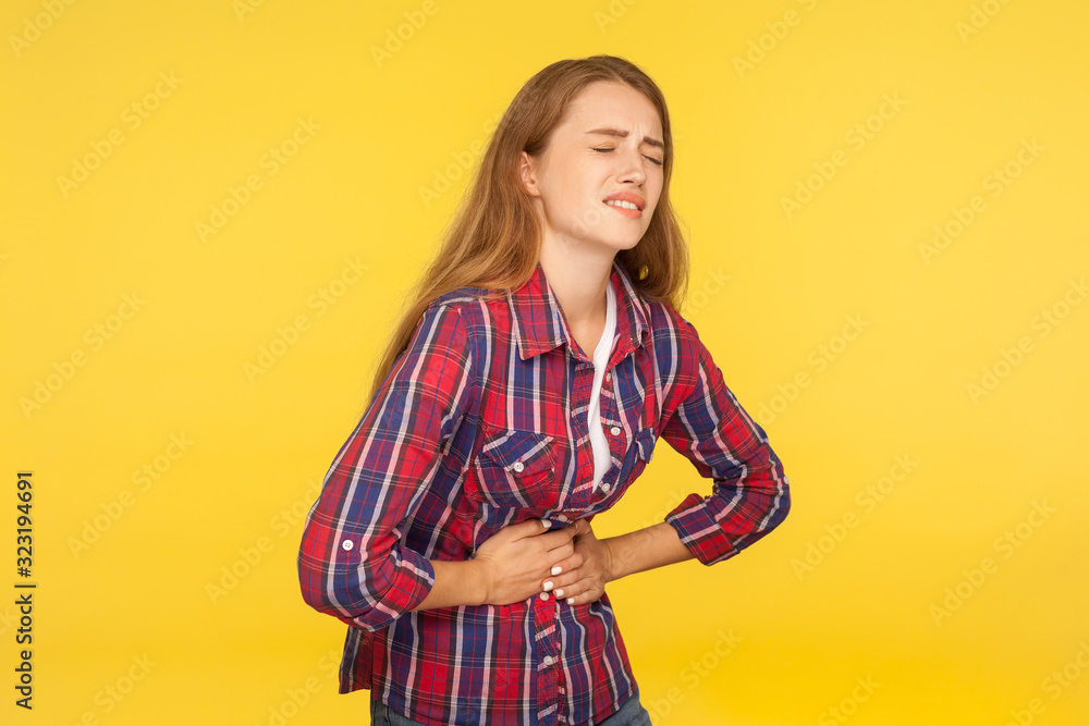 Abdominal pain. Portrait of unhealthy ginger girl in shirt clasping belly and suffering stomachache, indigestion symptoms, gastrointestinal disease. indoor studio shot isolated on yellow background Abdominal pain. Portrait of unhealthy ginger girl in shirt clasping belly and suffering stomachache, indigestion symptoms, gastrointestinal disease. indoor studio shot isolated on yellow background