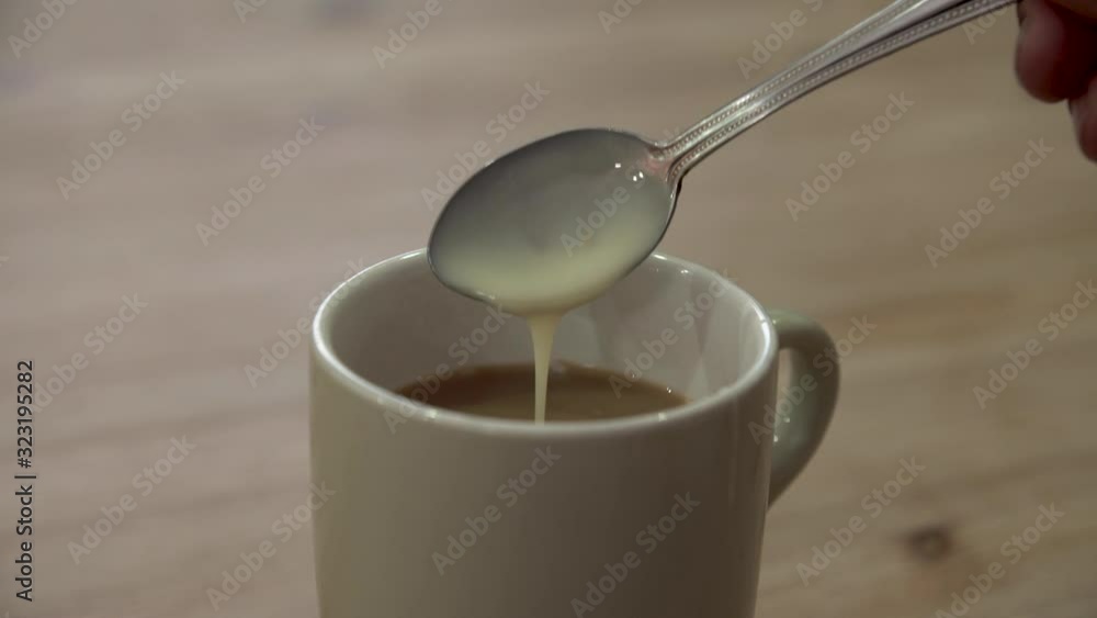 Hand pouring a tablespoon of condensed milk into a mug of coffee on a wooden table. Stirring a beverage with a spoon. Hand pouring a tablespoon of condensed milk into a mug of coffee on a wooden table. Stirring a beverage with a spoon.