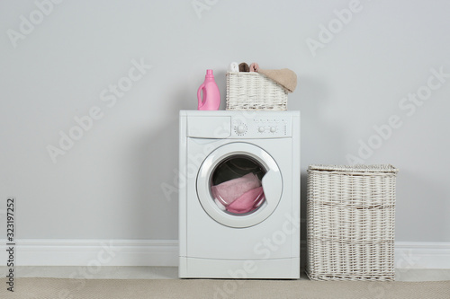 Modern washing machine with laundry, wicker baskets and detergent near white wall