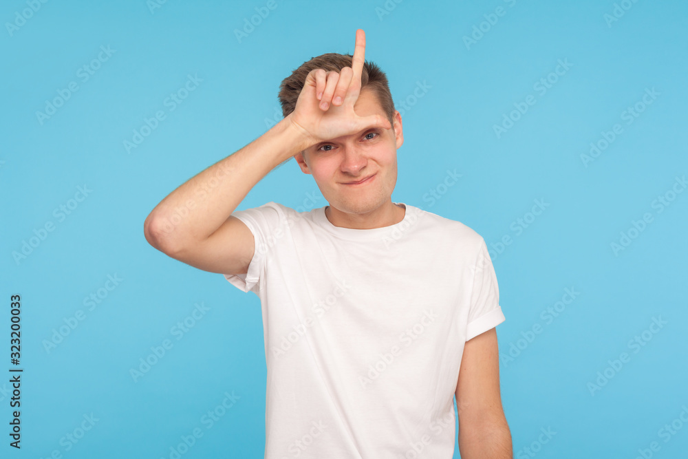 Portrait of upset depressed man in white t-shirt showing L sign on forehead, gesturing I'm loser, feeling defeated and blaming himself for failure. indoor studio shot isolated on blue background Portrait of upset depressed man in white t-shirt showing L sign on forehead, gesturing I'm loser, feeling defeated and blaming himself for failure. indoor studio shot isolated on blue background