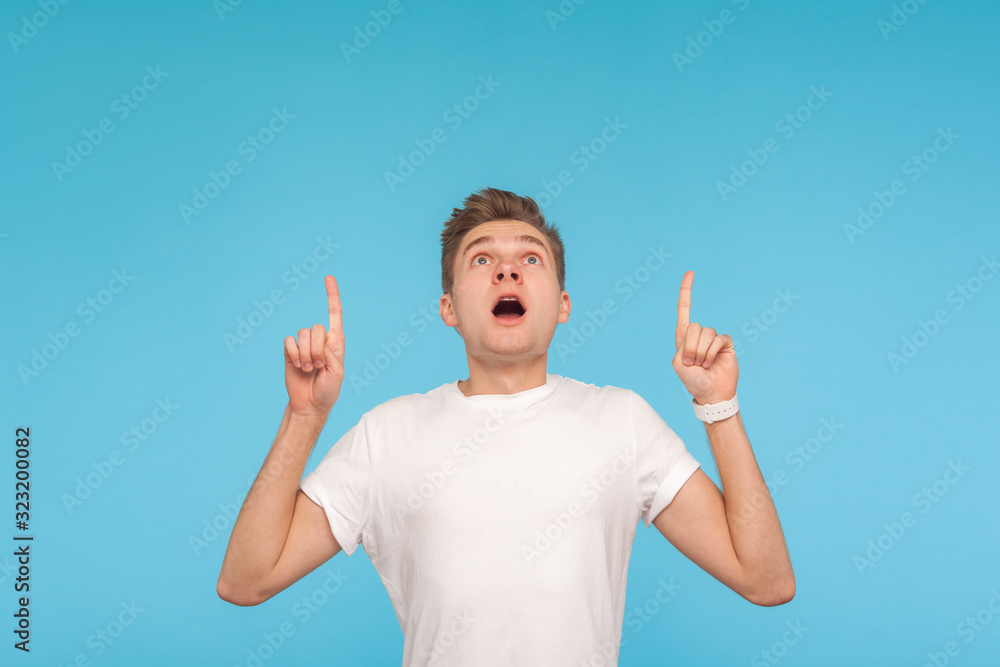 Shocking advertise here! Portrait of young man looking amazed and astonished while pointing up, showing blank copy space above head for crazy idea presentation. studio shot isolated on blue background Shocking advertise here! Portrait of young man looking amazed and astonished while pointing up, showing blank copy space above head for crazy idea presentation. studio shot isolated on blue background