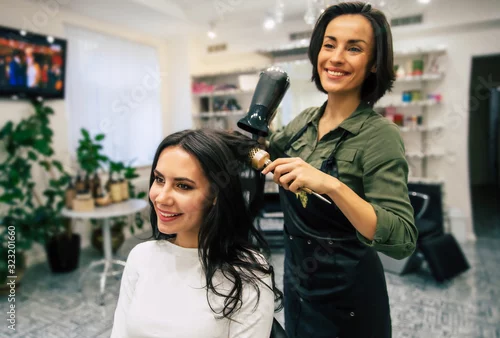 Fototapeta na wymiar Pure art. Young pretty woman is sitting in the armchair in a beauty salon getting her hair dried by a smiling professional stylist.