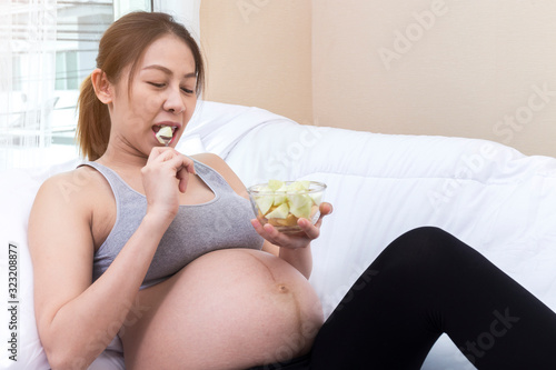 Happy Young Asian woman holding Bowl with delicious fruit salad  breakfast on sofa After she cooking doing fresh green salad eating many different vegetables during pregnancy healthy pregnancy concept