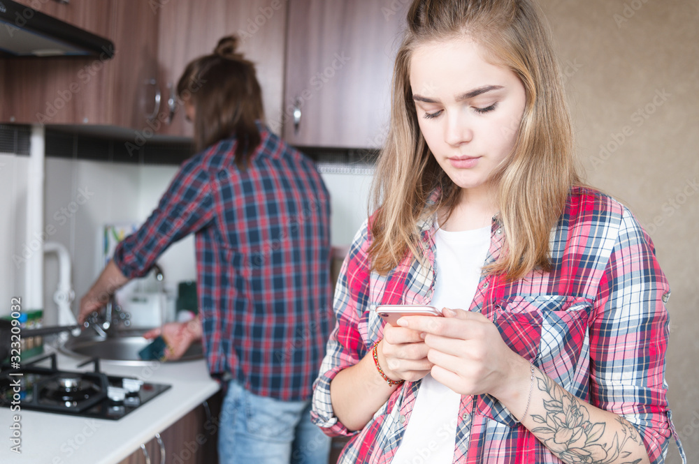 Young married couple in the kitchen. A girl with a tattoo writes a message on her mobile phone to her lover on the background of her husband who washes the dishes. Young married couple in the kitchen. A girl with a tattoo writes a message on her mobile phone to her lover on the background of her husband who washes the dishes.
