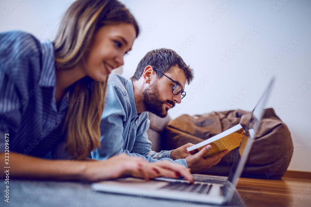Side view of handsome caucasian bearded man with eyeglasses reading book while his girlfriend smiling, lying next to him and using laptop. Selective focus on man. Side view of handsome caucasian bearded man with eyeglasses reading book while his girlfriend smiling, lying next to him and using laptop. Selective focus on man.