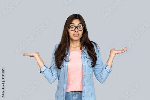 Unsure confused young adult indian woman standing isolated on grey background copy space. Uncertain doubtful hindu girl client customer doubting choosing looking at camera shrugging shoulders