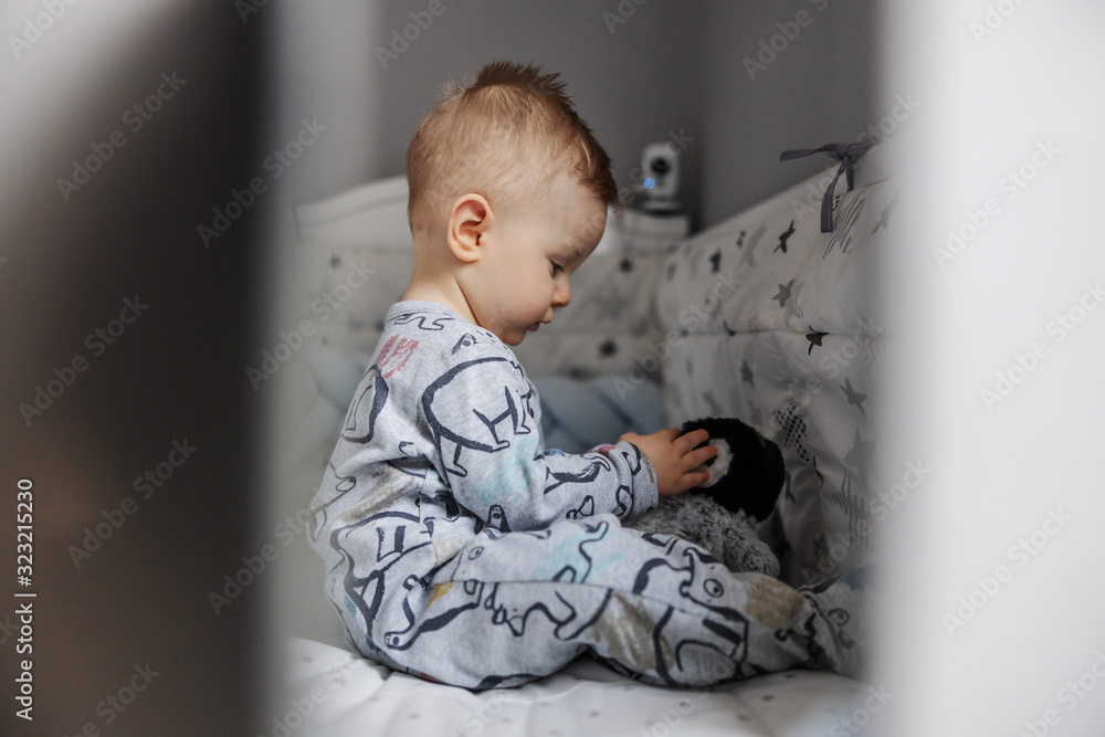 Fototapeta premium Portrait of charming blond little baby boy sitting in his crib and playing with his favorite penguin toy. Morning time.