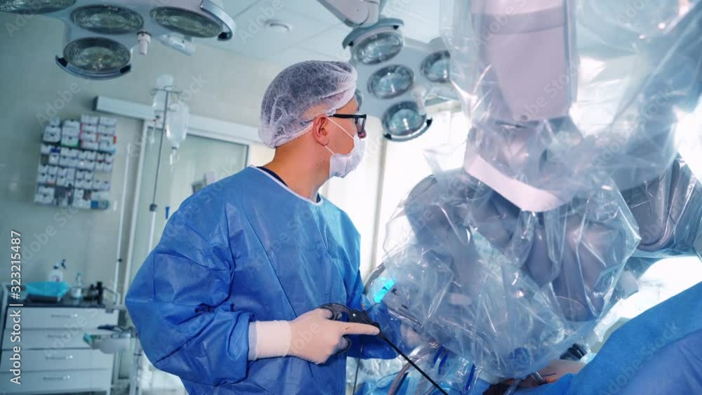 Medical specialist near the robotic device. Surgeon in blue protective uniform controls the work of medical robot on the screen. Medical specialist near the robotic device. Surgeon in blue protective uniform controls the work of medical robot on the screen.