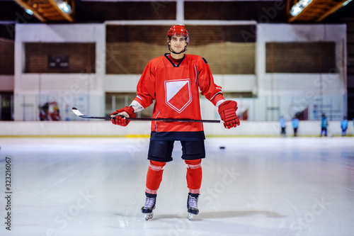 Full length of handsome hockey player holding stick while standing on ice in hall and looking at camera.