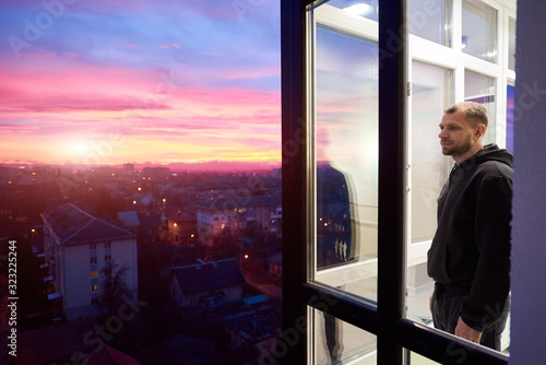 Side view of a man standing on a balcony with panoramic windows and looking into the distance while enjoying magical sunset. Urban landscape from height and a multi-colored fiery sky with setting sun