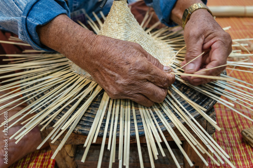 Hands of old artisan craftsman elderly working weaving rattan and bamboo to make ancient handmade handcraft wicker traditional Thai wooden hat photo