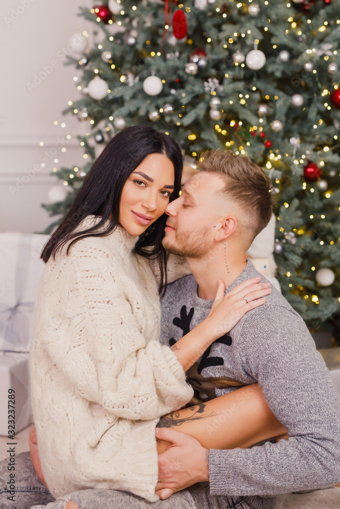 young beautiful couple, brunette girl in a white sweater, a blond man in a gray sweater sitting near a Christmas tree young beautiful couple, brunette girl in a white sweater, a blond man in a gray sweater sitting near a Christmas tree