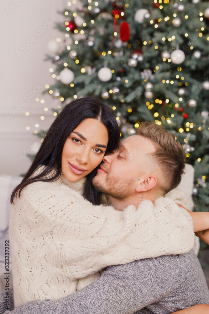young beautiful couple, brunette girl in a white sweater, a blond man in a gray sweater sitting near a Christmas tree young beautiful couple, brunette girl in a white sweater, a blond man in a gray sweater sitting near a Christmas tree