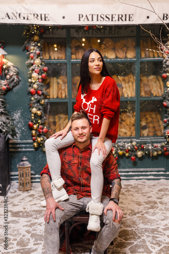 Young beautiful couple, a brunette girl in a red sweater and white pants, a man in a red shirt are sitting on the streets of Paris against the background of a window of a bakery shop