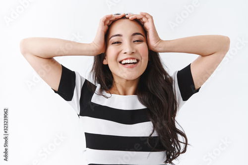 Carefree, upbeat happy asian girl with relaxed delighted smile, holding hands on head and grinning, being in good mood, feeling relieve or happiness, standing white background