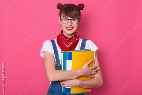 Close up portrait of happy good looking student with colorful paper folder, wears bandana, t shirt and overalls, looks at camera and smiling, has funny hairstyle, poses isolated over rosy background.