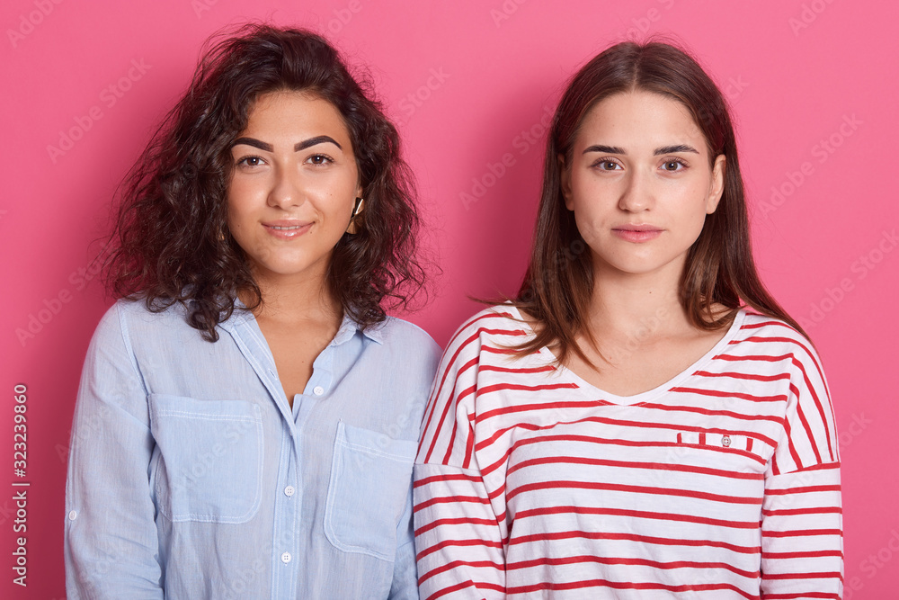 Horizontal shot of two females with calm face. Women standing isolated over pink stidio background, one lady wearing blue blouse, another striped shirt, looking directly at camera. People concept. Horizontal shot of two females with calm face. Women standing isolated over pink stidio background, one lady wearing blue blouse, another striped shirt, looking directly at camera. People concept.