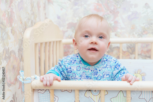 A little boy, a child with blue eyes is standing in a crib and smiling at home. Learns to stand at the support. Portrait, close-up