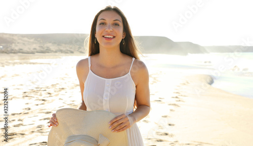 Portrait of beautiful smiling carefree woman walking on the beach in white dress holding straw hat looking at camera with overexposed background. Copy space. photo