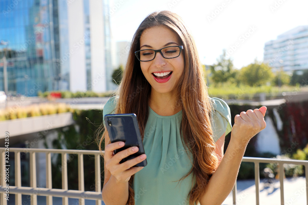 Excited young business woman receiving good news on mobile phone celebrating with fist up in modern city district in spring time