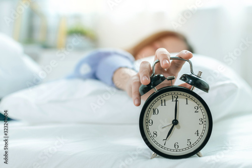 young asian woman sleeping on bed pressing snooze button on black vintage alarm clock at seven o'clock morning in bed room at home, lifestyle, good morning, healthy sleep and joyful weekend concept