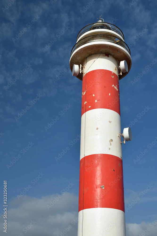 Toston lighthouse, El Cotillo, Fuerteventura, Spain. Close up of exterior with red and white stripes, peeling paint. Blue sky and small clouds.