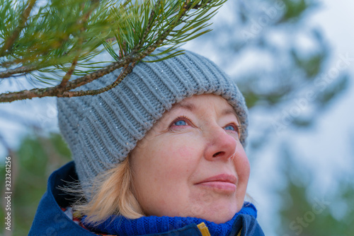 Female wearing blue knitted hat. Cheerful mature woman smiling. Outdoors cold weather portrait. Face close up. Pine tree background