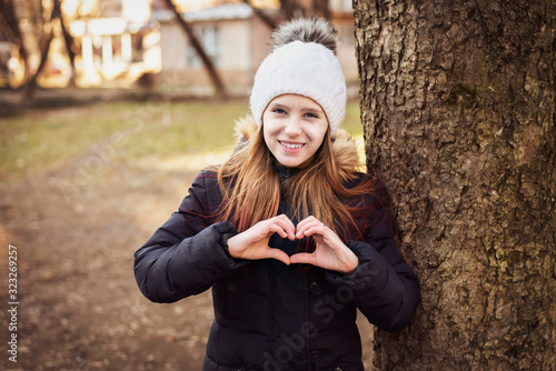 The young girl shows heart with her hands, she is waiting for the spring which she loves very much. Girl dressed in warm winter clothes. Welcome spring