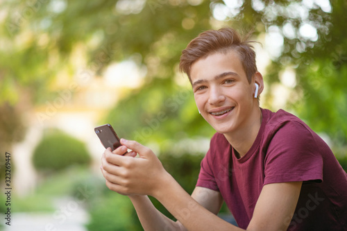Happy teenage boy is using mobile phone, outdoors. Close-up portrait of a smiling young man with smartphone, in park. Cheerful teenager in casual clothes with cell phone in park. Soft focus