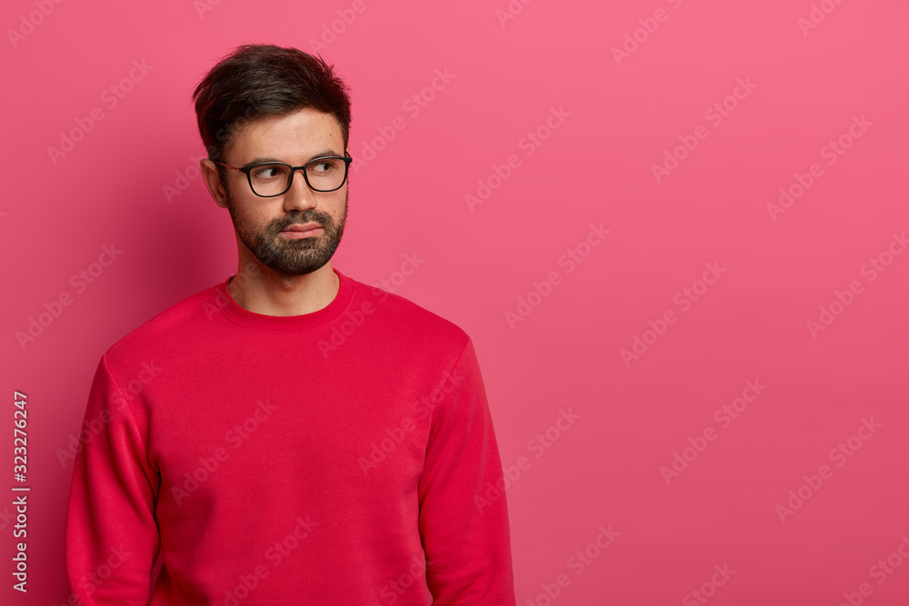 Horizontal shot of serious looking bearded man looks aside with thoughtful expression, thinks over new exclusive service, wears spectacles and red jumper, considers good sale offer, isolated on pink Horizontal shot of serious looking bearded man looks aside with thoughtful expression, thinks over new exclusive service, wears spectacles and red jumper, considers good sale offer, isolated on pink