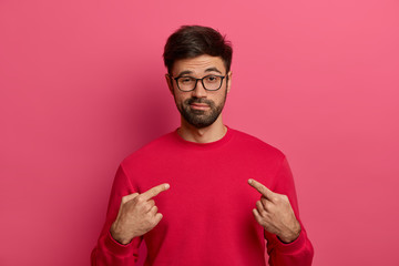 I can handle it. Assertive determined man with beard points at himeself, feels proud and brags about own achievements, proposes own help, wears red sweater, eyewear, stands against pink background