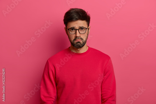 Dejected sad man with thick beard, has unlucky day, faces problems in life, looks desperately and gloomy, purses lips, wants to cry, wears red sweater, isolated on pink background. Negative feelings