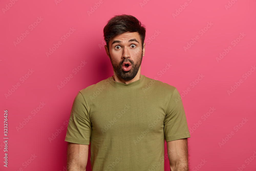 Embarrassed bearded guy reacts on false information, keeps mouth opened, looks at camera with astonishment and disbelief, wears casual clothes, poses indoor. Young man receives shocking news