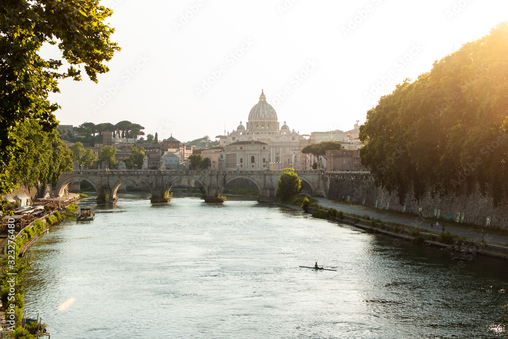Young woman looking at St Peter's basilica from a bridge on tiber river at sunet, taking pictures with smartphone. Panoramic view cityscape of Rome, Italy.