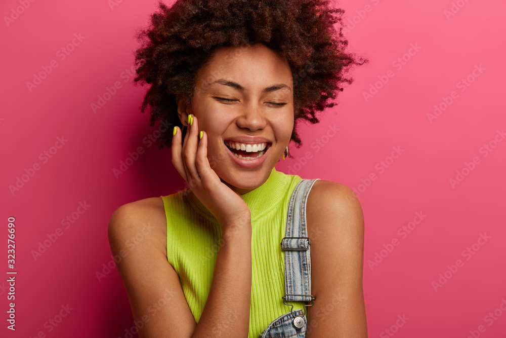 Headshot of optimistic curly haired woman laughs out loudly, smiles toothily, shows white teeth, dressed in fashionable clothes, giggles with eyes closed, models over pink background. Emotions concept Headshot of optimistic curly haired woman laughs out loudly, smiles toothily, shows white teeth, dressed in fashionable clothes, giggles with eyes closed, models over pink background. Emotions concept
