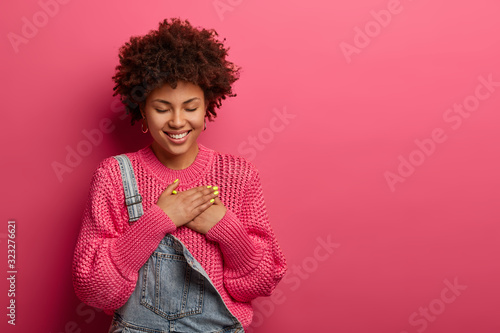 Lovely cheerful Afro American woman presses palms to heart, has grateful expression, thankful for received gift, closes eyes, wears fashionable clothes, isolated on pink wall, blank space aside photo