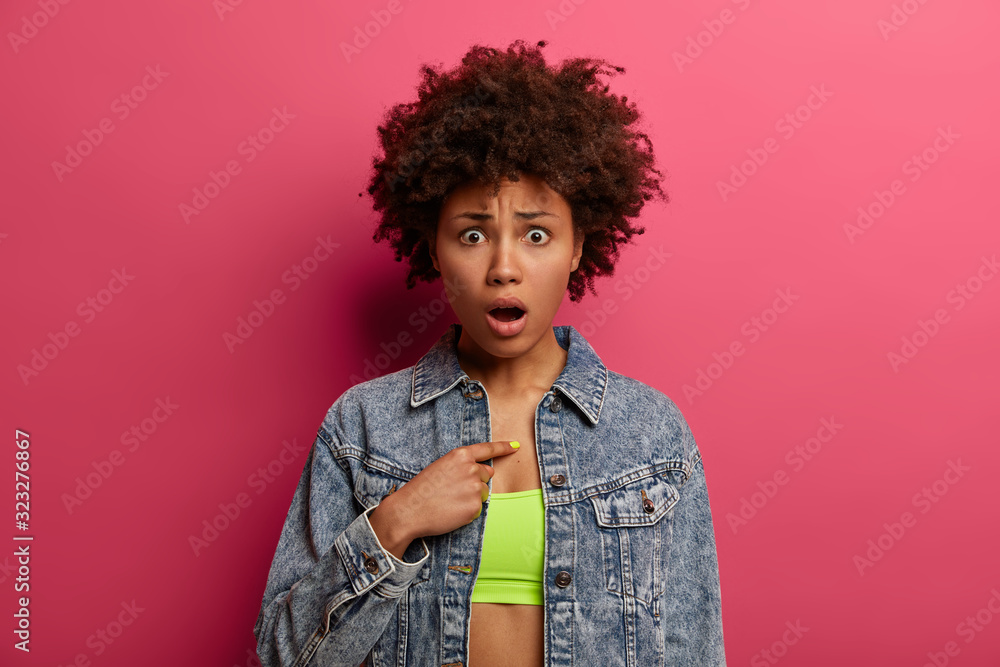 Embarrassed young Afro American woman points at herself, asks question, doesnt expect being chosen, keeps mouth opened, wears top and denim jacket, has disappointed face expression, poses indoor Embarrassed young Afro American woman points at herself, asks question, doesnt expect being chosen, keeps mouth opened, wears top and denim jacket, has disappointed face expression, poses indoor