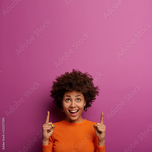 Pleased attractive young woman has nice idea, points above on blank space, has broad smile, curious expression, advertises new object, wears orange jumper, isolated over purple wall, empty space