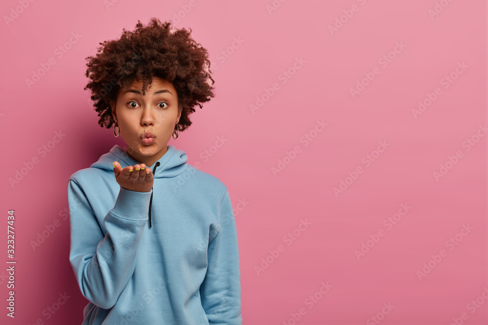 Lovely curly haired ethnic woman sends air kiss, pulls palm towards mouth, wears blue sweatshirt, looks directly at camera, expresses love to husband, poses over rosy background, has flirty expression Lovely curly haired ethnic woman sends air kiss, pulls palm towards mouth, wears blue sweatshirt, looks directly at camera, expresses love to husband, poses over rosy background, has flirty expression