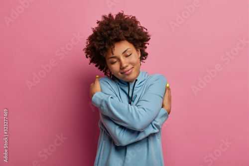 Coziness and self esteem concept. Pleased dark skinned woman with Afro hair, embraces herself, feels comfort in new sweatshirt, tilts head and smiles gently, feels fullfilled, isolated over rosy wall