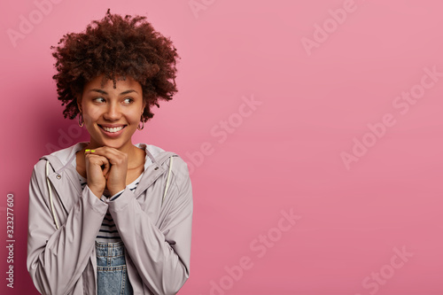 Horizontal shot of cute Afro American woman keeps hands under chin, sees beautiful view aside, daydreams and thinks about something pleasant, wears casual windbreaker, looks at something cute