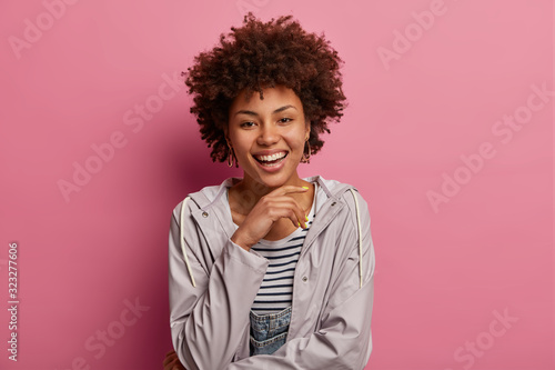 Lovely optimistic woman with Afro hairstyle, wears casual anorak, touches chin gently, listens good news with pleasure, has carefree expression, isolated over rosy pastel wall, poses relaxed