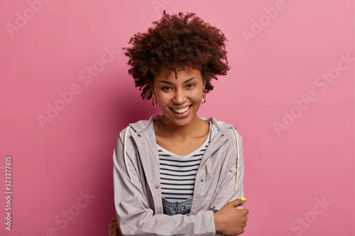Happy cute woman with timid calm face expression, crosses hands over body, looks with gentle smile at camera, wears casual windbreaker, expresses positive emotions, isolated on pink background