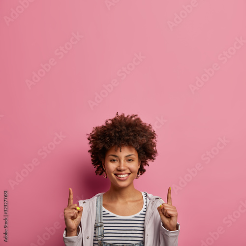 Here what you need. Satisfied Afro American woman points above, shows copy space, smiles happily, dressed casually, isolated over pink pastel background, advertises item for you. People and promotion