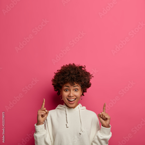 Look, advertise here. Happy delighted woman with curly Afro hair, points above, smiles cheerfully, tries to grab your attention, shows empty place for your promotional text, wears white hoodie